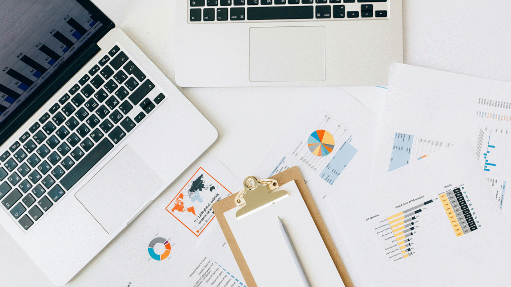 A desk cluttered with open notebooks, scattered data pages featuring graphs and charts, and a laptop displaying analytical information, symbolizing in-depth financial analysis and strategic planning in business economics.