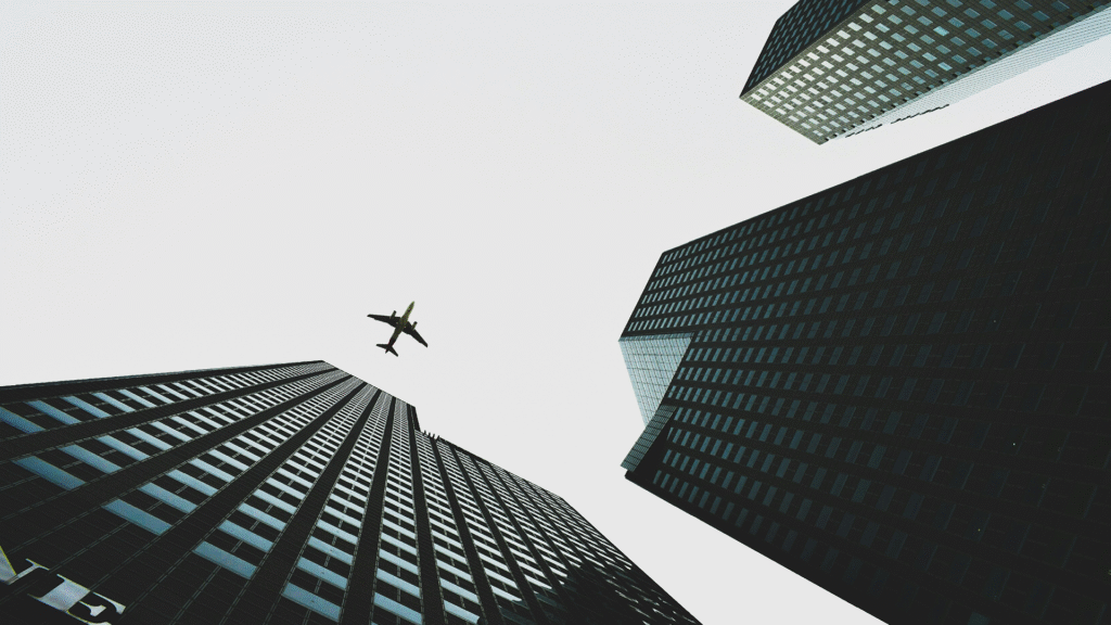 An airplane in flight against a bright sky, with the tops of two to three tall, modern buildings visible from below, symbolizing the global scale and commanding presence of Fortune 500 corporations.