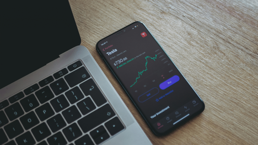 A laptop and a smartphone display fluctuating stock charts, specifically showing a Tesla share graph, illustrating real-time investment monitoring and the psychology of investing in volatile markets.