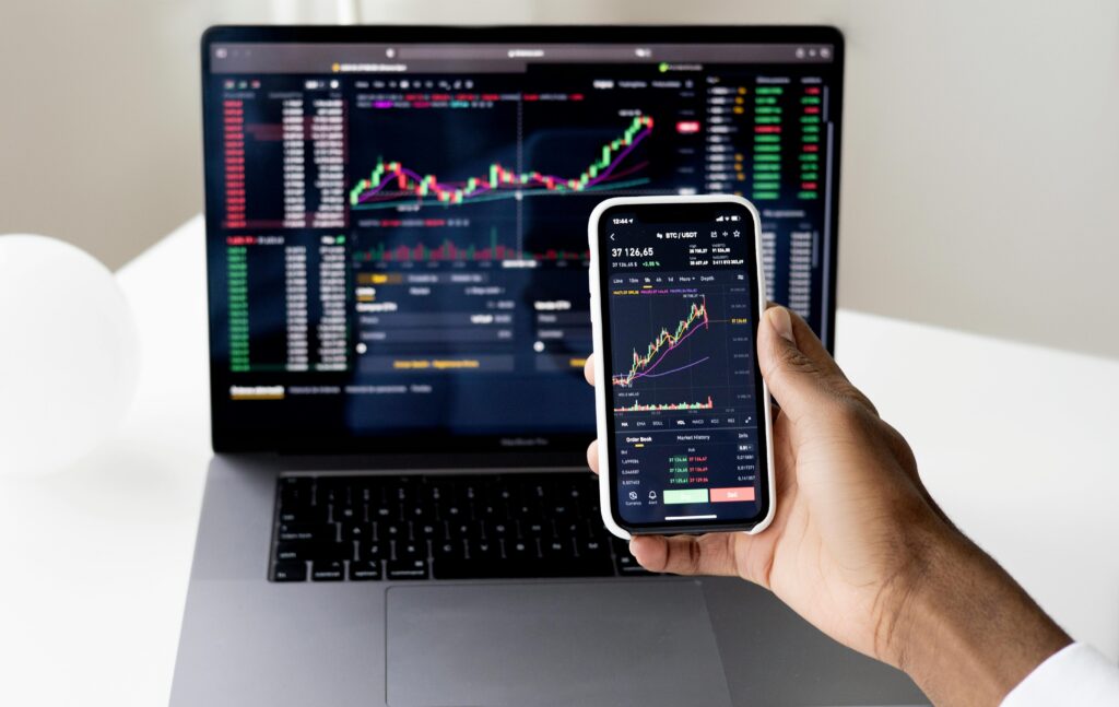 A person holding a smartphone displaying stock market charts, with a laptop showing similar stock trends in the background.