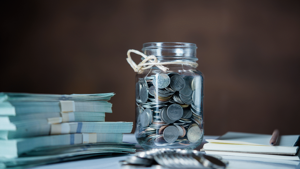 A clear glass bottle filled with coins, placed beside several bundles of cash, symbolizing a well-prepared emergency fund.