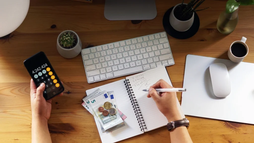 Person practicing money management by using a calculator, taking notes in a notebook, analyzing financial graphs, and counting cash on a desk.
