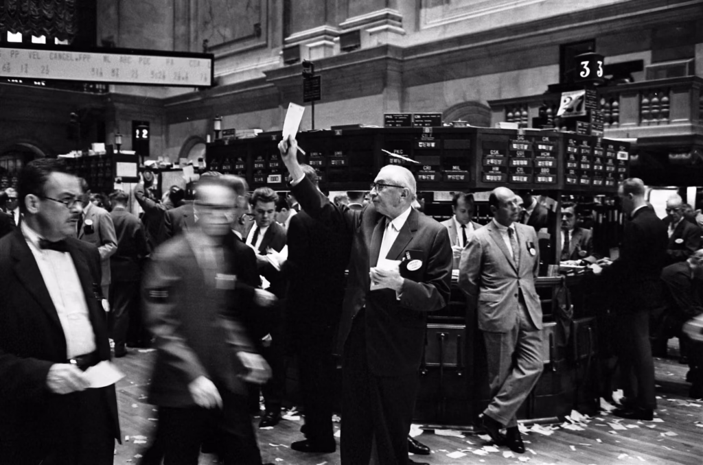 Historic black and white photo of New York Stock Exchange trading floor with stock traders in suits shouting orders, waving papers, and executing trades before electronic trading - showing how stock markets worked traditionally