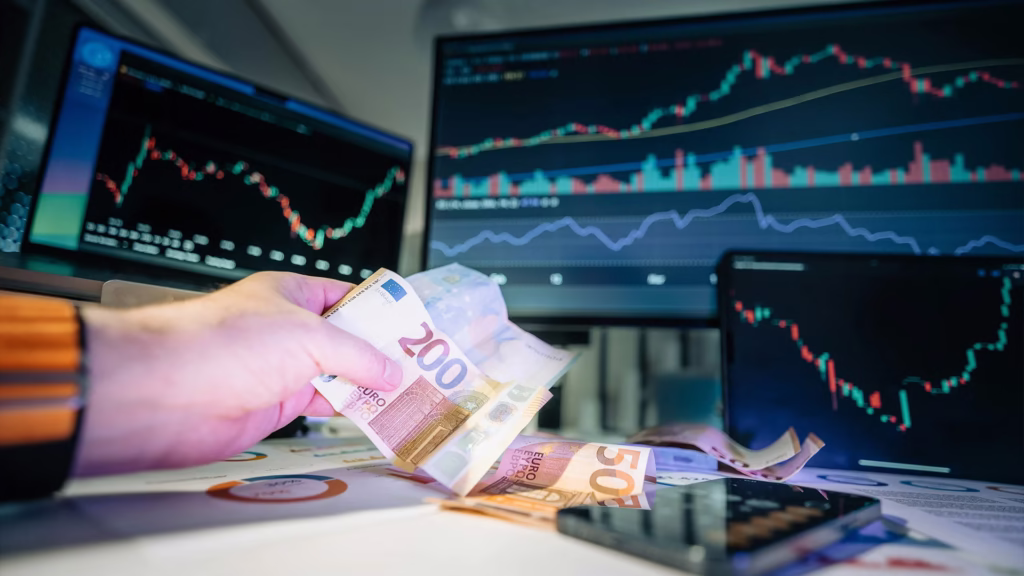 Investor holding Euro currency cash while monitoring multiple computer screens displaying stock market charts with candlestick patterns and volume indicators - representing investment strategies, portfolio management, and strategic money allocation for beginners