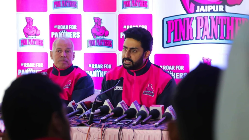 Abhishek Bachchan at Jaipur Pink Panthers press conference wearing team jersey speaking with media representing his Pro Kabaddi League franchise ownership worth over 200 crore rupees making him one of India's most successful celebrity sports team owners alongside coach