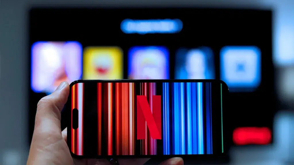 Hand holding smartphone displaying Netflix logo with TV screen showing Netflix content in background