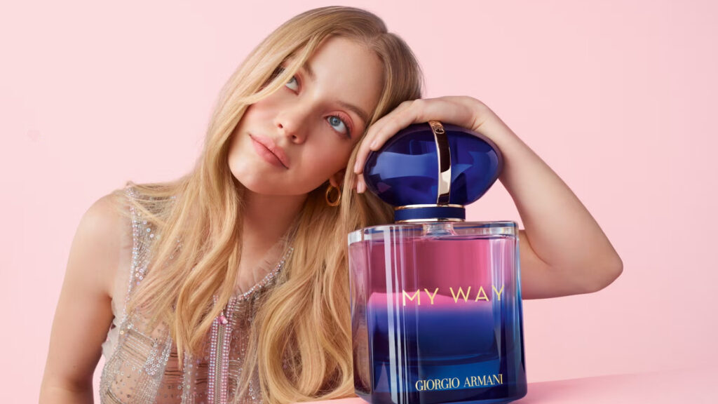Sydney Sweeney holding Armani Beauty perfume bottle in pink background showcasing global beauty ambassador partnership