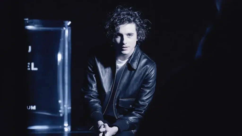 Timothée Chalamet in black leather jacket seated in dark studio next to illuminated Bleu de Chanel fragrance bottle during campaign shoot