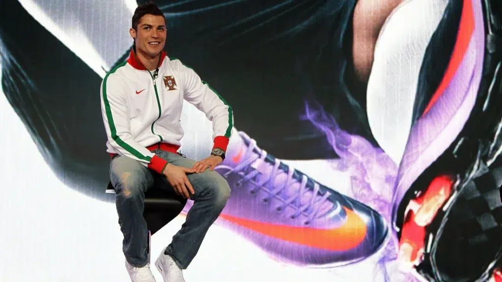 Cristiano Ronaldo smiling in Portugal white tracksuit seated on stage at Nike Mercurial football boot launch event with large boot campaign display behind him